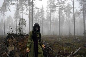 A green clad figure wearing a plague doctors mask standing in a foggy forest.